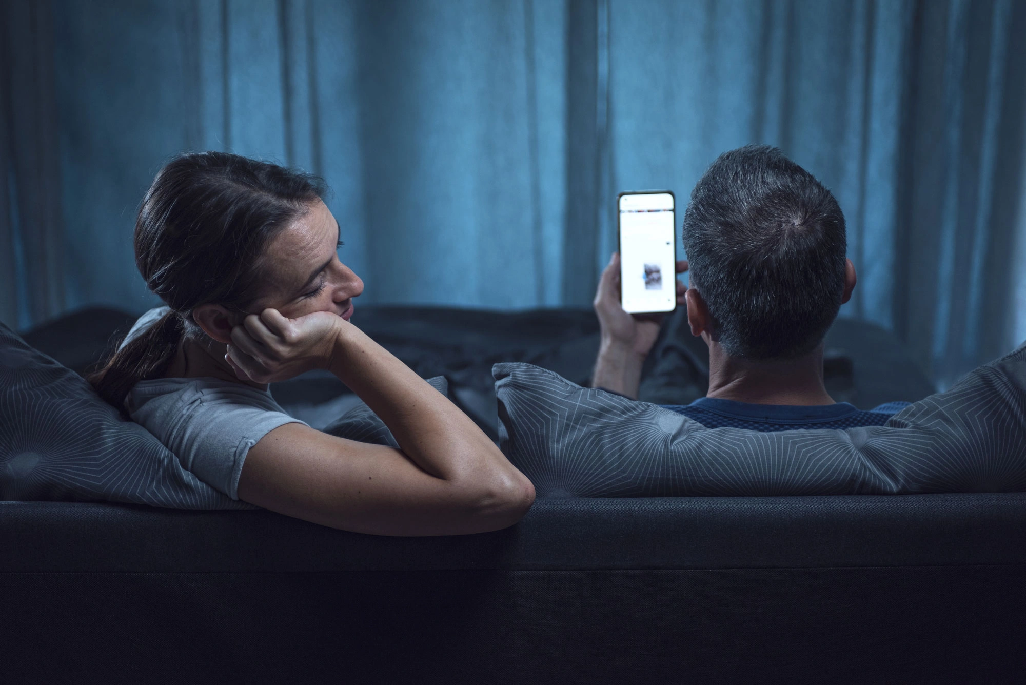 Para leży wieczorem w łóżku, a mężczyzna przegląda telefon. Zdjęcie symbolizuje ciągłe scrollowanie, przebodźcowanie cyfrowe i wpływ smartfonów na zdrowie psychiczne.