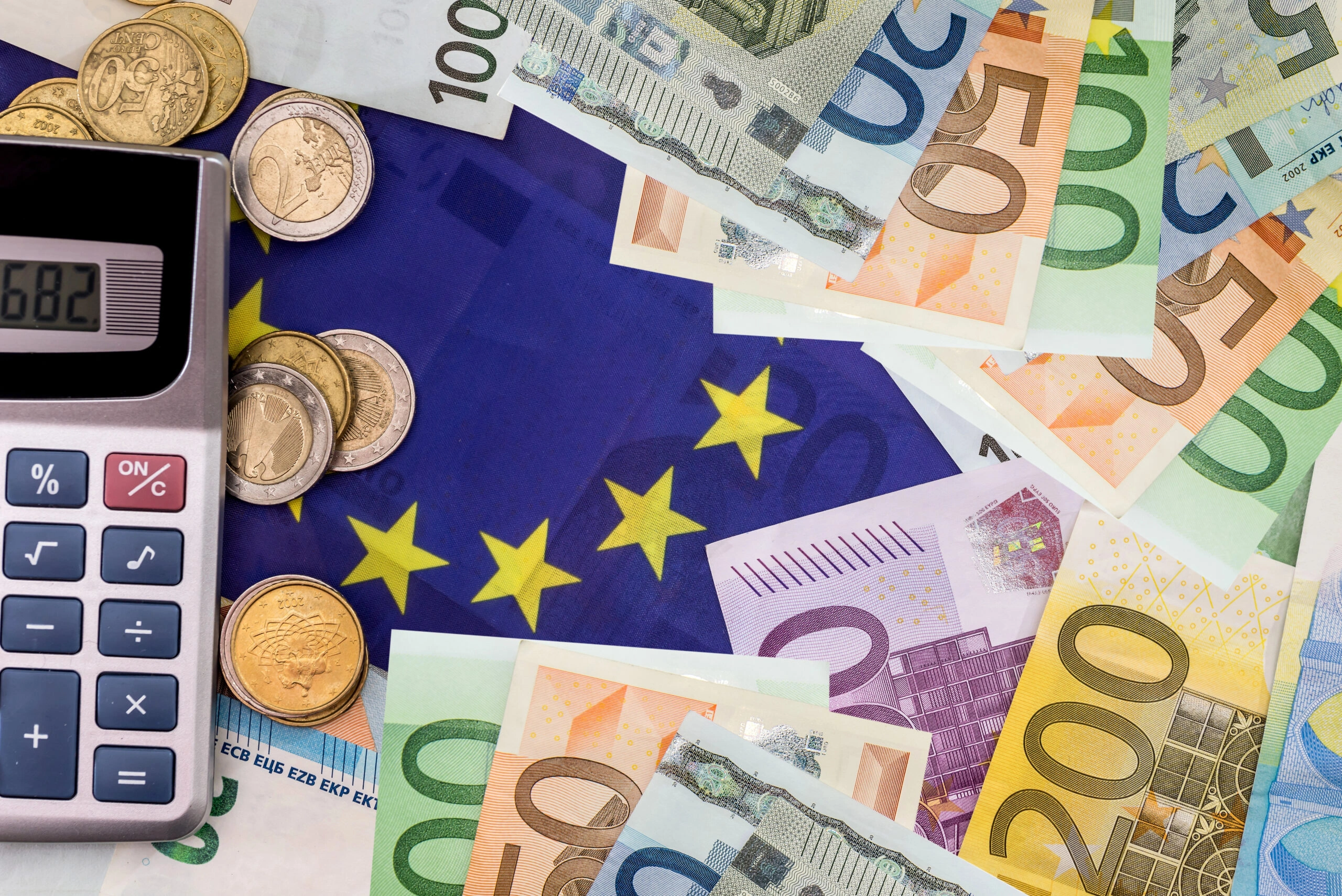 Alt: Kalkulator, monety i banknoty euro na tle flagi Unii Europejskiej symbolizują koszty profilaktyki i leczenia uzależnień.