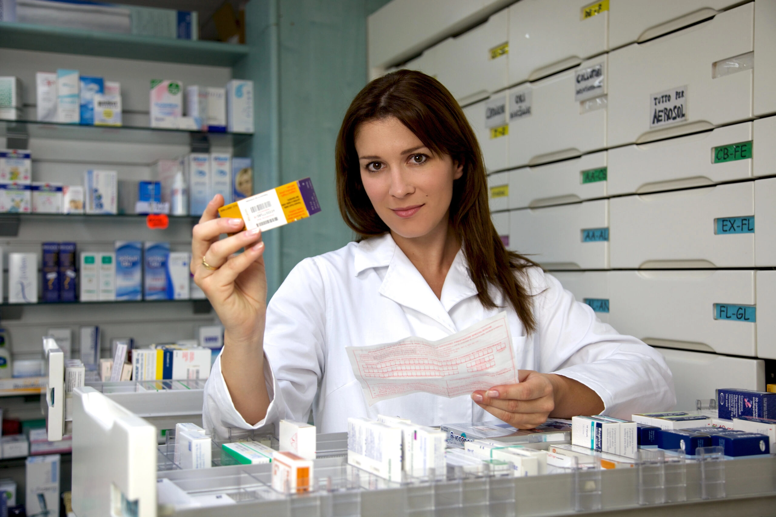Farmaceutka w aptece trzyma opakowanie leku i receptę, symbolizując dostęp do leków na receptę oraz kontrolę ich wydawania.